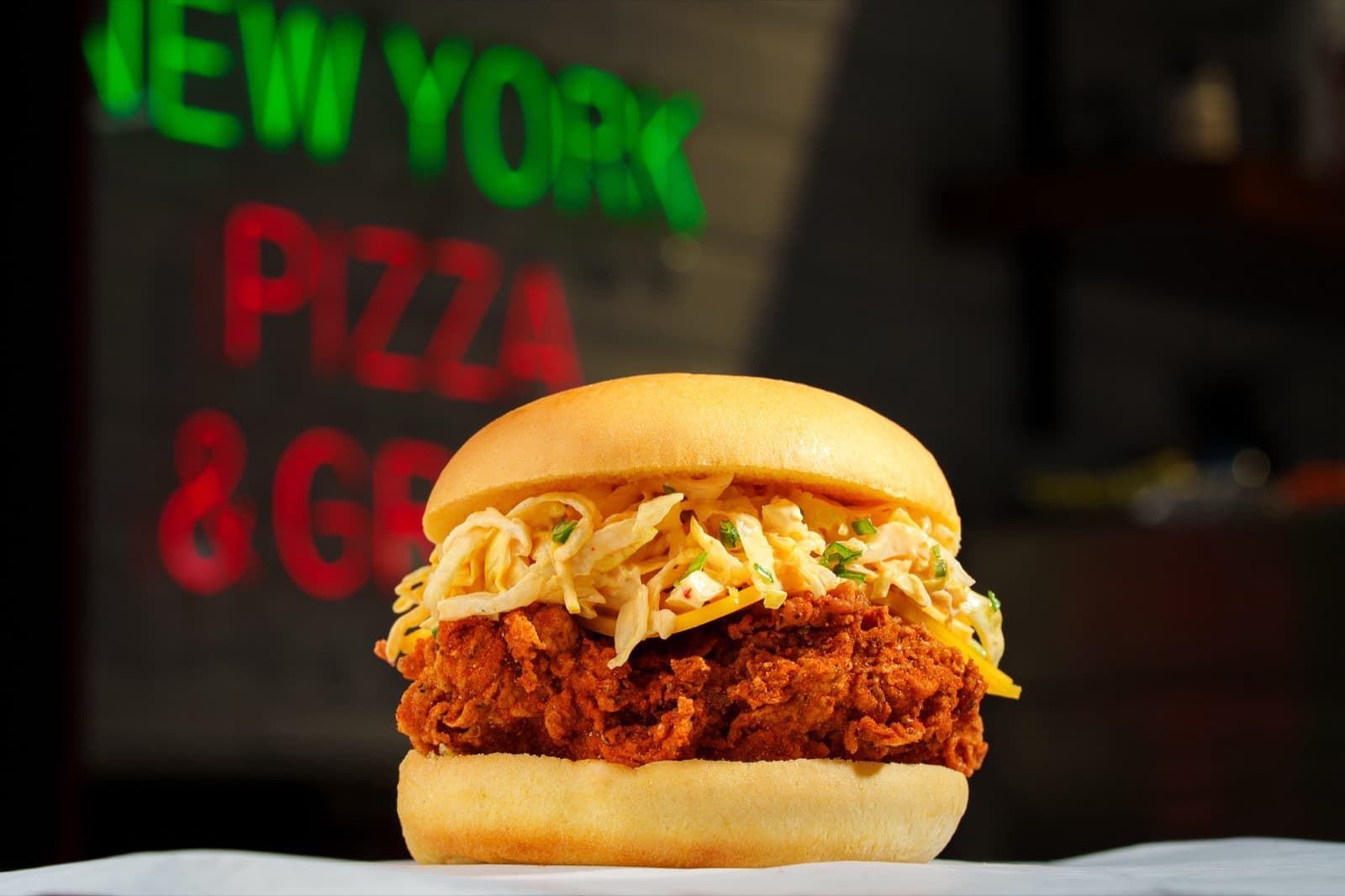 Burger au poulet croustillant et coleslaw devant l’enseigne néon New York Pizza & Grill